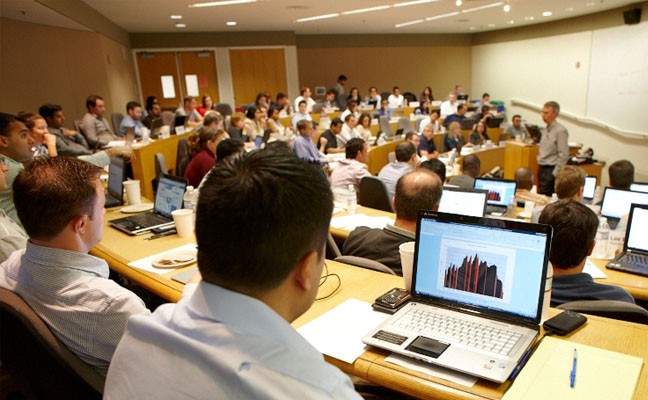 Students in a lecture hall listen to a speaker while referencing information on laptops.