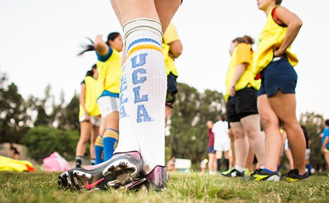 Close-up of woman wearing knee-high UCLA athletic socks and soccer cleats, with other women in sports gear in the background