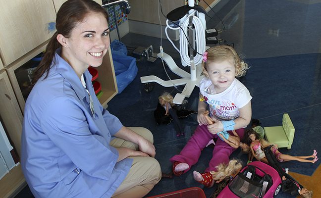 medical staff member sits with toddler girl playing with dolls