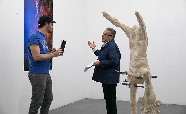 Graduate student standing and listening to professor in white art gallery space, standing near painting and sculpture.