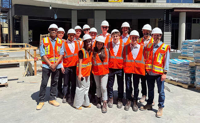 Students with orange construction vests and white helmets stand together in a construction site.