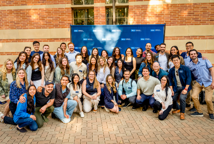 A large group of UCLA Anderson students smiling and posing together outdoors in front of a UCLA Anderson branded backdrop, reflecting the warmth and diversity of the Anderson community.