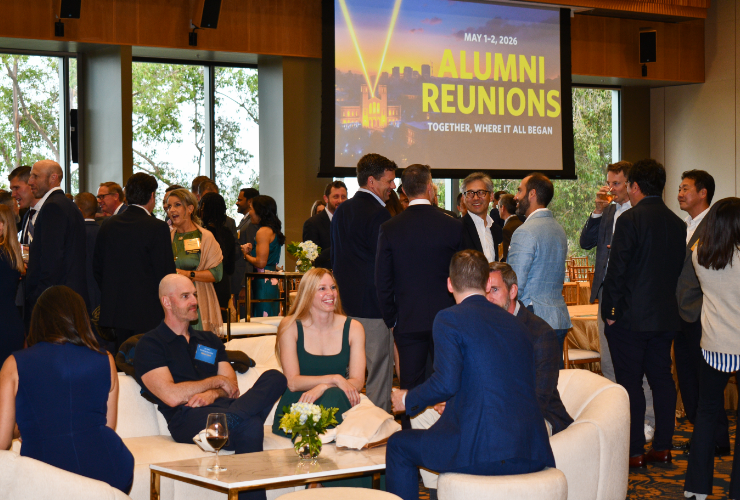 UCLA Anderson alumni mingling and conversing at an alumni reunion reception, with an Alumni Reunions branded screen visible in the background.
