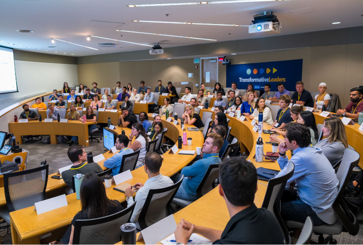 A full UCLA Anderson classroom of students seated and attentively engaged during an academic session, with a Transformative Leaders banner visible in the background.